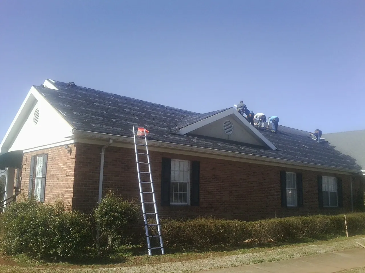 Crew tearing shingles off a brick ranch roof with a ladder set — Metal Roof Repair work by our crew in Hanson